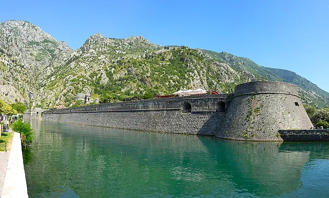 Die Stadtmauer von Kotor am Fluss Škurda, mit grünem Wasser und dem Berghang dahinter