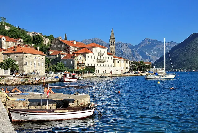 Die Uferpromenade von Perast mit Steinhäusern, roten Dächern und einem Glockenturm, Boote auf dem Wasser