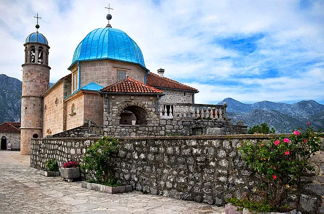 Die Kirche Unserer Lieben Frau vom Felsen mit blauer Kuppel auf ihrem Inselchen, hinter einer Steinmauer mit Rosen