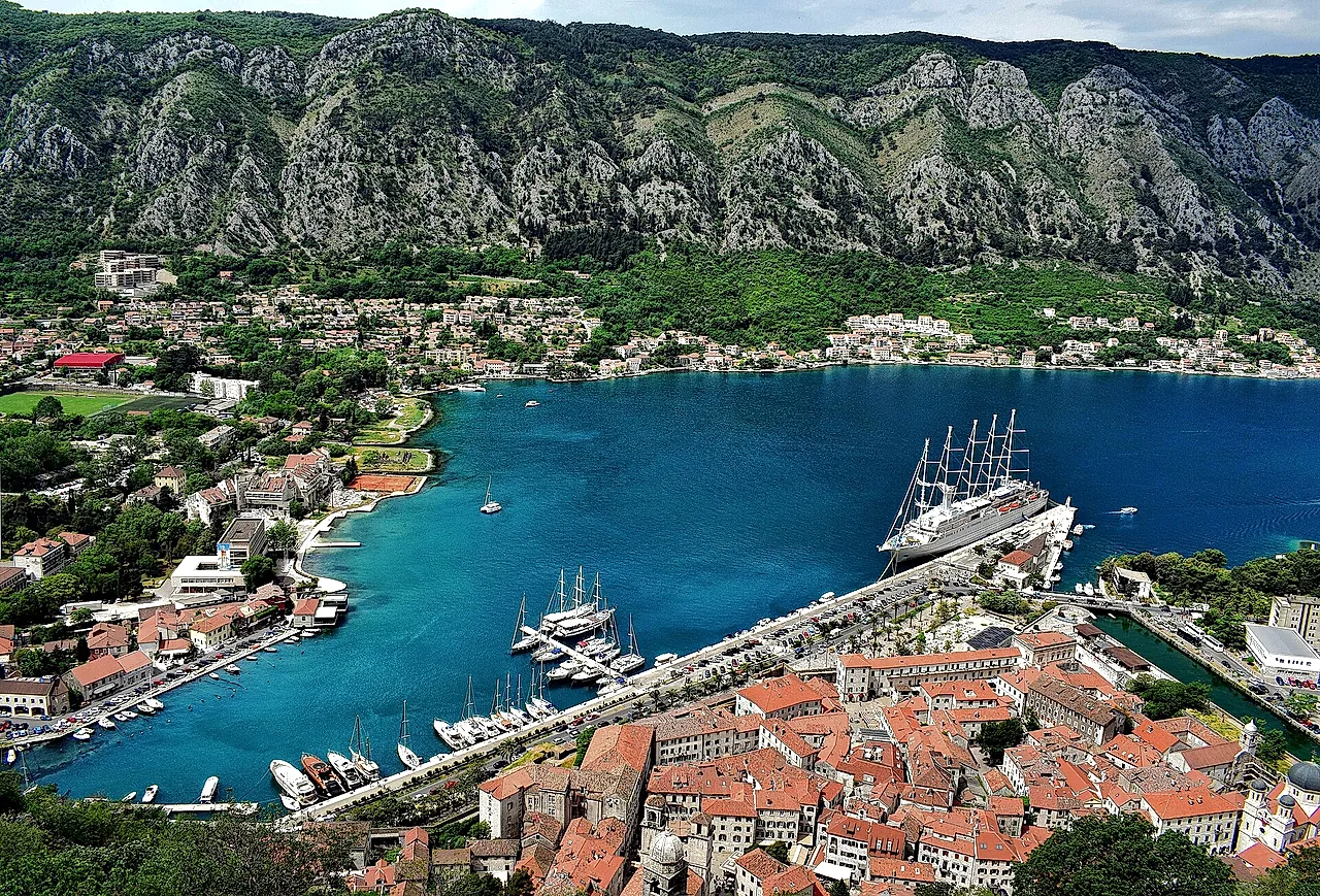 Die Altstadt von Kotor mit roten Dächern an der blauen Bucht, ein Großsegler liegt am Kai, dahinter Berge